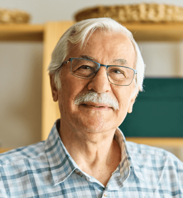 Smiling senior man with white hair, thick mustache, and blue-framed glasses, wearing a light blue plaid button-up shirt, standing in a cozy workshop or office setting with wooden shelves and baskets in the blurred background