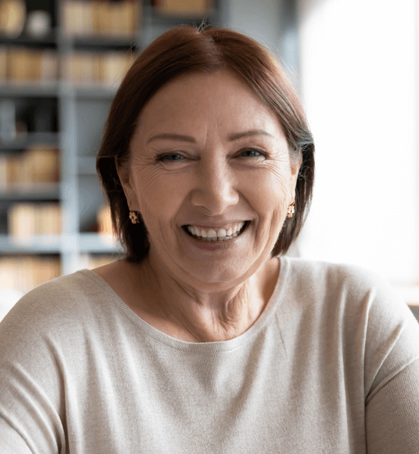 Smiling mature woman with shoulder-length auburn hair, warm expression, subtle laugh lines, wearing small gold stud earrings and a light beige crew-neck sweater, standing in front of a blurred bookshelf background