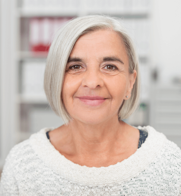 Smiling mature woman with short silver-gray bob haircut, warm brown eyes, subtle smile lines, wearing small stud earrings and a textured white knit sweater, standing in an office setting with blurred bookshelves and red binders in the background